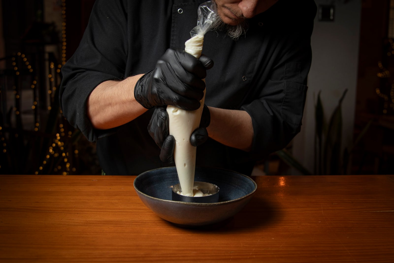 Un chef con guantes negros rellenando masa en un molde con manga pastelera, sobre un plato en una mesa de madera.